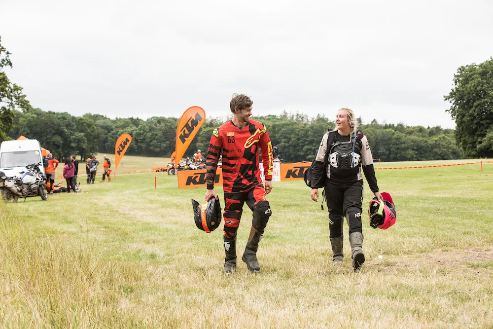 A man and a woman walk in a field carrying their motorcycle helmets
